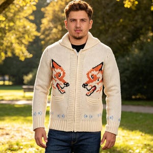 May include: Cream-coloured zip-up cardigan sweater with two orange fox head designs on the front. The sweater has a shawl collar, two pockets, and blue floral accents on the cuffs. The person is wearing blue jeans.