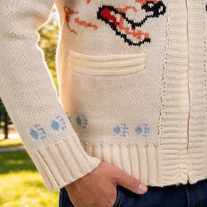 May include: Cream-coloured zip-up cardigan sweater with a textured knit. The sweater features a design with red, black, and orange accents. The cuffs have blue paw print details. The sweater has two front pockets.