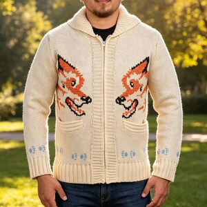 May include: Cream-coloured zip-up sweater with a shawl collar and two front pockets. The sweater features a fox design in orange and black on the chest. Light blue paw prints are on the cuffs. The sweater is made of a thick knit material.