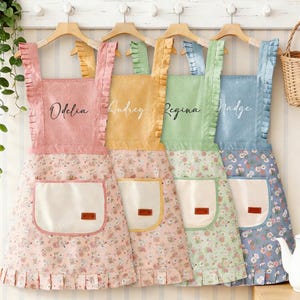 May include: Four colourful aprons in pink, yellow, green, and blue, each with a floral pattern on the skirt and a contrasting pocket. The aprons are hanging on wooden hangers, with names like "Odelia" and "Audrey" written on them.