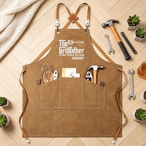 May include: A brown canvas apron with leather straps and pockets, featuring the text "The Grillfather" and a name. The image includes tools, small succulents in pots, and a glass of amber liquid, suggesting a grilling theme.