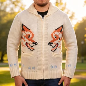 May include: Cream-coloured zip-up cardigan with two orange and black fox face designs. The sweater features a shawl collar, two front pockets, and blue paw print details on the cuffs. A warm knitwear garment for cooler weather.