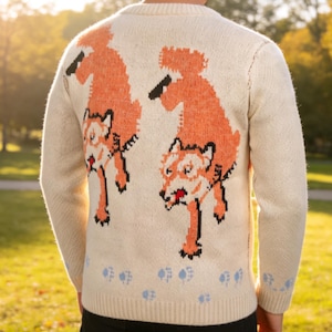 May include: Cream-coloured jumper featuring a repeating orange fox design on the back. The foxes are depicted in a pixelated style, with black and white accents. Blue paw prints are at the bottom of the jumper.
