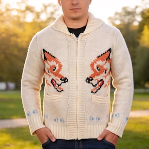 May include: Cream-coloured zip-up cardigan sweater with fox face designs in orange and black on the front. The sweater has a shawl collar, two front pockets, and blue paw prints on the cuffs.