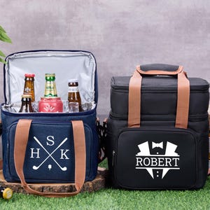 Peut inclure: Deux sacs isothermes, un bleu marine et un noir, avec des poignées et des sangles marron. Le sac bleu marine a un monogramme sur le thème du golf, et le sac noir présente un motif de smoking avec le nom "ROBERT". Les deux sacs sont ouverts, révélant des boissons.