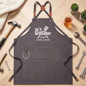 Puede incluir: Un delantal gris carbón con detalles de cuero y el texto "The GrillFather 2026 LOVE, DAVID". El delantal tiene bolsillos y un anillo de metal. Herramientas y plantas rodean el delantal, incluyendo un martillo, llaves y suculentas.