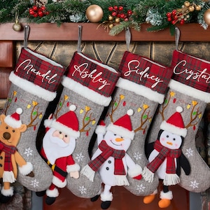 May include: Four personalised Christmas stockings hang from a mantelpiece. Each stocking is grey with a red plaid cuff and features a different plush character: a reindeer, Santa, a snowman, and a penguin. Names are embroidered on the cuffs.