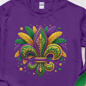 May include: A purple sweatshirt featuring a detailed fleur-de-lis design in green, gold, and purple, surrounded by feather accents. The design is centered on the front of the sweatshirt. A Mardi Gras mask and hat are visible.