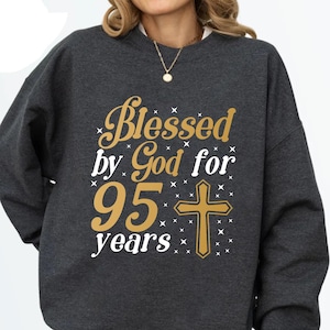 May include: A dark gray sweatshirt with the text "Blessed by God for 95 years" in gold and white lettering. A gold cross is centered below the text.