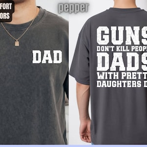 May include: Dark grey t-shirt with the word "DAD" printed in white on the front. The back of the shirt features the text "GUNS DON'T KILL PEOPLE DADS WITH PRETTY DAUGHTERS DO" in white. The shirt is part of the "Comfort Colors" collection.