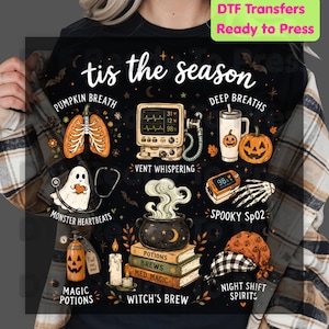 May include: A black t-shirt featuring a graphic print with the text 'tis the season' and various medical-themed Halloween illustrations including lungs, a heart monitor, a ghost with a stethoscope, a pumpkin, a pulse oximeter, and a cauldron.