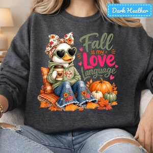May include: A dark heather gray sweatshirt featuring a graphic of a duck wearing a floral bow, heart-shaped sunglasses, and a green hoodie. The duck holds a coffee cup and wears blue jeans. Text reads Fall is my Love Language with pumpkins.