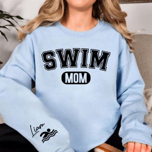 May include: Light blue sweatshirt with black text that reads "SWIM MOM" and a black oval with the word "MOM" inside. The left sleeve has a black swimmer icon and the text "Liam" in cursive.