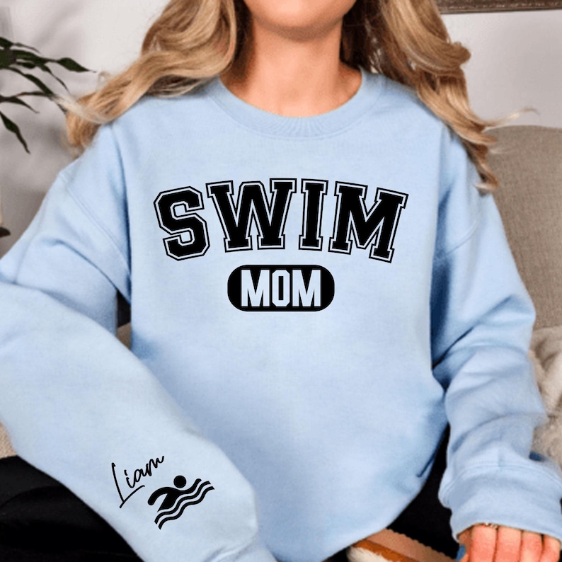 May include: Light blue sweatshirt with black text that reads "SWIM MOM" and a black oval with the word "MOM" inside. The left sleeve has a black swimmer icon and the text "Liam" in cursive.