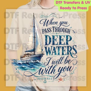 May include: A beige t-shirt featuring a graphic of a sailboat on blue ocean waves. The text reads: When you PASS THROUGH DEEP WATERS I will be with you. ISAIAH 43:2. The design includes a small seashell and a starfish at the bottom.