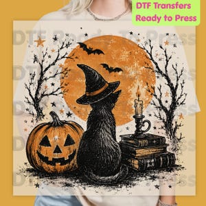 May include: A cream-colored t-shirt featuring a graphic print of a black cat wearing a witch hat, sitting beside a jack-o'-lantern, a stack of books, and a lit candle. The background shows a large orange moon, bare tree silhouettes, and bats.