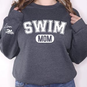May include: A dark gray sweatshirt with the text "SWIM MOM" in white. The text "SWIM" is in a large, bold font, and the text "MOM" is in a smaller, oval-shaped font. The sweatshirt also has a small swimmer icon on the left sleeve.