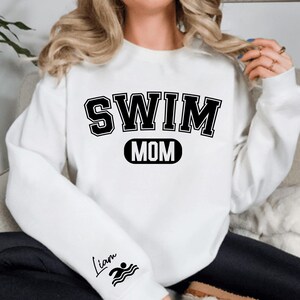 May include: White sweatshirt with black text that reads "SWIM MOM" in a varsity style font. The left sleeve has a small black graphic of a swimmer and the name "Liam" written in cursive.