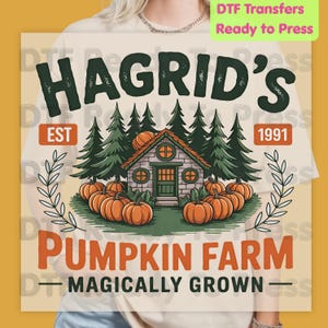 May include: A graphic print featuring a stone cabin surrounded by pine trees and pumpkins. The text reads HAGRID'S EST 1991 PUMPKIN FARM MAGICALLY GROWN. The design uses dark green, orange, and grey tones on a light-colored t-shirt background.