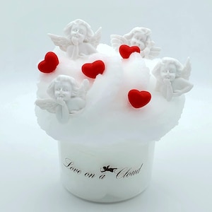 May include: White ceramic candle jar with a white cotton cloud on top. The cloud is decorated with five small white cherubs and five red hearts. The jar has a black label that reads "Love on a Cloud".