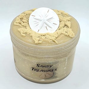 Puede incluir: Un recipiente de pl&aacute;stico transparente lleno de una sustancia de color beige, coronado con un d&oacute;lar de arena blanco y detalles beige texturizados. El recipiente tiene una etiqueta que dice "Sandy Treasures".