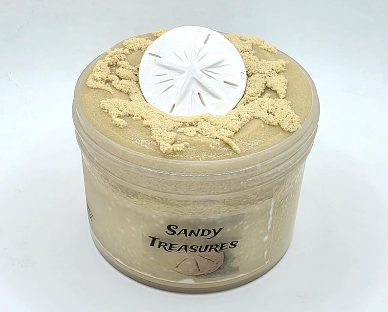 Puede incluir: Un recipiente de pl&aacute;stico transparente lleno de una sustancia de color beige, coronado con un d&oacute;lar de arena blanco y detalles beige texturizados. El recipiente tiene una etiqueta que dice "Sandy Treasures".