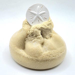 Puede incluir: Una escultura beige y redondeada con una textura similar a la arena, coronada por un d&oacute;lar de arena blanco. El d&oacute;lar de arena tiene un dise&ntilde;o en forma de estrella. La escultura evoca un tema de playa y decoraci&oacute;n costera.