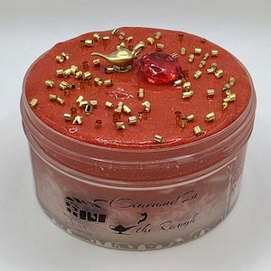 May include: A clear plastic jar filled with red textured slime, topped with small gold cylindrical beads, a gold-colored genie lamp charm, and a red faceted gem. The label on the side features a graphic of a treasure chest and lamp with the text A Diamond In the Rough.