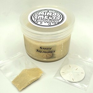Puede incluir: Un recipiente de pl&aacute;stico transparente lleno de arena de color beige, etiquetado como "Sandy Treasures". Acompa&ntilde;ado de una peque&ntilde;a bolsa de arena y un d&oacute;lar de arena en una bolsa de pl&aacute;stico transparente. El recipiente tiene una etiqueta blanca con el texto "Mind Melts Emporium."