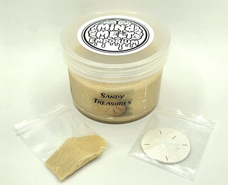 Puede incluir: Un recipiente de pl&aacute;stico transparente lleno de arena de color beige, etiquetado como "Sandy Treasures". Acompa&ntilde;ado de una peque&ntilde;a bolsa de arena y un d&oacute;lar de arena en una bolsa de pl&aacute;stico transparente. El recipiente tiene una etiqueta blanca con el texto "Mind Melts Emporium."