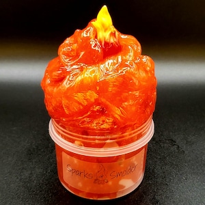 May include: Orange and yellow slime in a clear plastic container with a white lid. The container has a label that says "Sparks & Smolder".