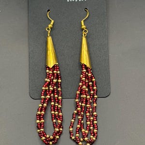 May include: Gold-tone metal and red beaded earrings. The earrings have a conical top and multiple strands of red and gold beads. The earrings are displayed on a black card that reads "Handmade with love."