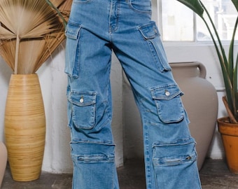 May include: Blue denim cargo pants featuring a high-waisted fit, a button and zipper fly closure, and multiple utility pockets on the thighs and shins. The pants have a wide-leg silhouette and are paired with a white cropped top and pink sneakers.