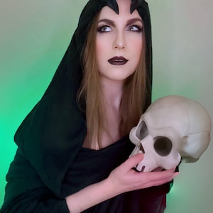 May include: A person wearing a black hooded cloak and a decorative headpiece with antler-like elements, holding a skull. The person has dark lipstick and is looking to the side. The background has a green gradient.