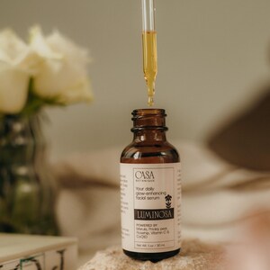 May include: A brown glass bottle of facial serum with a dropper dispensing the product. The label reads "CASA BOTANIQUE LUMINOSA" and "Your daily glow-enhancing facial serum". The bottle is on a light-colored rock. Net Wt. 1 oz / 30 mL.