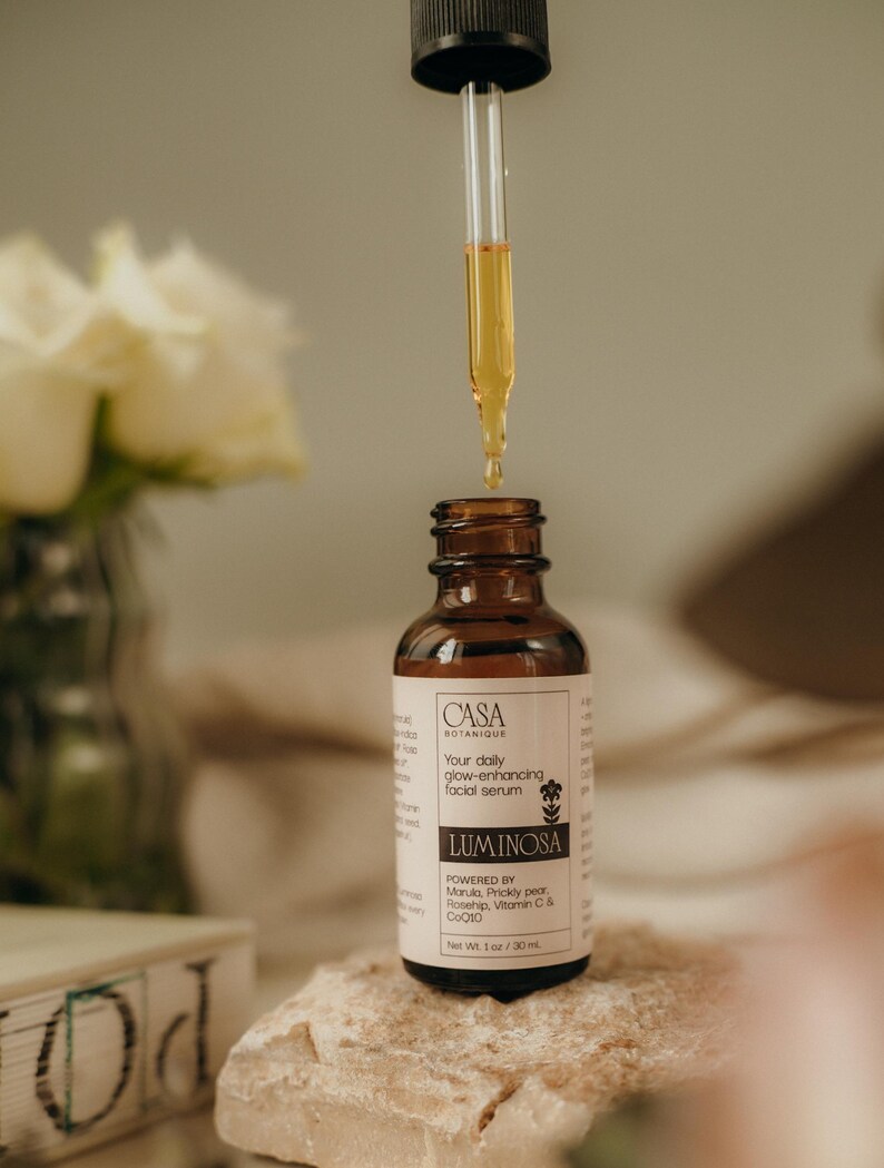 May include: A brown glass bottle of facial serum with a dropper dispensing the product. The label reads "CASA BOTANIQUE LUMINOSA" and "Your daily glow-enhancing facial serum". The bottle is on a light-colored rock. Net Wt. 1 oz / 30 mL.