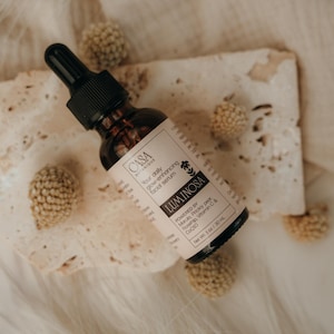 May include: A brown glass bottle of facial serum with a black dropper, labeled "Luminosa." The label is light pink with black text. The bottle rests on a piece of textured paper, surrounded by small, round, beige decorative elements.