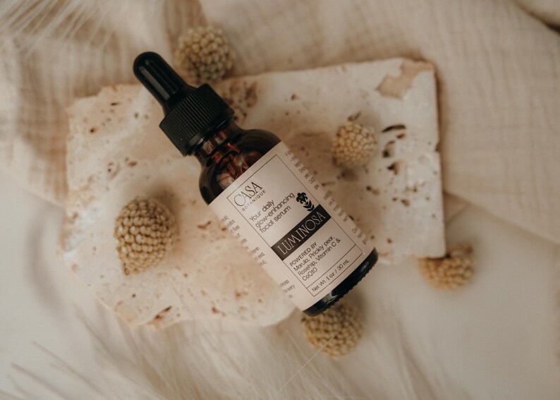 May include: A brown glass bottle of facial serum with a black dropper, labeled "Luminosa." The label is light pink with black text. The bottle rests on a piece of textured paper, surrounded by small, round, beige decorative elements.