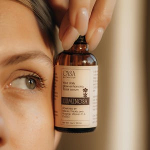 May include: A brown glass bottle of facial serum, held by a hand, with the brand name "CASA BOTANIQUE" and the product name "LUMINOSA" visible. The label states the serum is powered by Marula, Prickly pear, Rosehip, Vitamin C & CoQ10. Net Wt. 1 oz / 30 ml.
