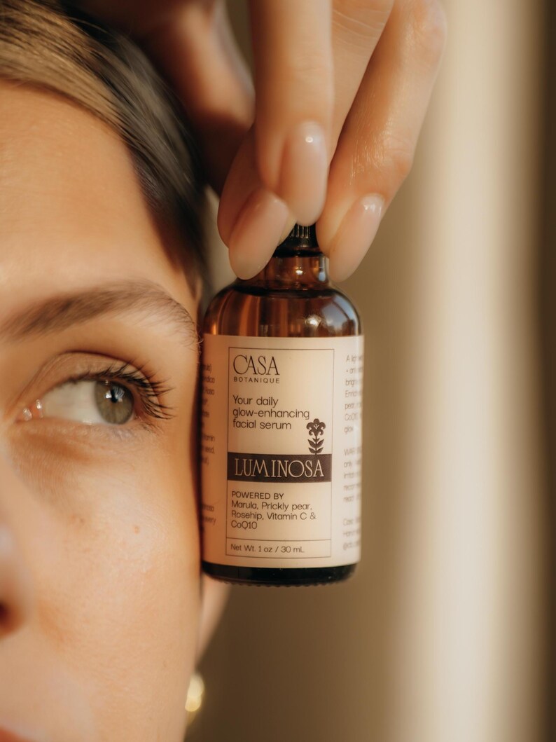 May include: A brown glass bottle of facial serum, held by a hand, with the brand name "CASA BOTANIQUE" and the product name "LUMINOSA" visible. The label states the serum is powered by Marula, Prickly pear, Rosehip, Vitamin C & CoQ10. Net Wt. 1 oz / 30 ml.