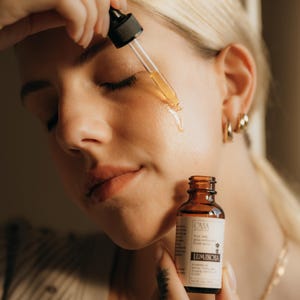 May include: A woman applying a clear serum from a dropper to her face. The serum is in a small brown glass bottle labeled "Luminosa". The woman has blonde hair and is wearing gold hoop earrings.