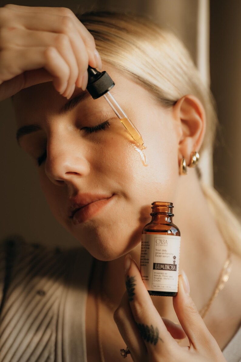 May include: A woman applying a clear serum from a dropper to her face. The serum is in a small brown glass bottle labeled "Luminosa". The woman has blonde hair and is wearing gold hoop earrings.