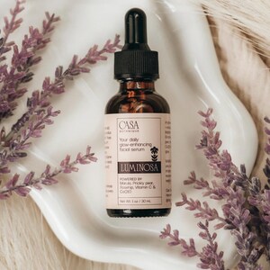 May include: A brown glass bottle of facial serum with a black dropper, labeled "Luminosa" and "Casa Botanique." The bottle is on a white, wavy dish, surrounded by purple lavender sprigs. The serum is 1 oz / 30 ml.