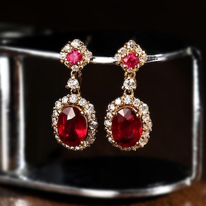Könnte beinhalten: Ein Paar goldene Ohrhänger mit ovalen, tiefroten Edelsteinen, die von kleinen Diamanten umgeben sind. Jeder Ohrring hat einen kleineren roten Edelstein an der Spitze, ebenfalls von Diamanten umgeben. Dunkler Hintergrund.