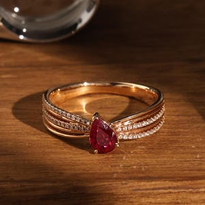 Puede incluir: Un anillo de oro con una piedra preciosa roja oscura en forma de pera. El anillo presenta múltiples bandas adornadas con pequeñas piedras brillantes. El anillo está sobre una superficie de madera, con un objeto borroso en el fondo.