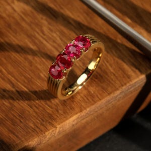 Puede incluir: Un anillo de banda de oro con cinco piedras preciosas de rubí de forma ovalada engastadas en una fila. El anillo está sobre una superficie de madera.