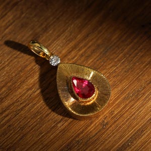 May include: A gold pendant with a teardrop-shaped design. The pendant features a vibrant red gemstone at its center, surrounded by a textured gold setting. A small, round diamond is set above the teardrop. The pendant is displayed on a wooden surface.