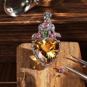 Peut inclure: Un pendentif en argent avec une grande pierre jaune triangulaire à la base, surmonté d'une monture en argent texturé ornée de deux pierres roses rondes, une pierre verte centrale et deux petites pierres lavande, avec des pinces à proximité.