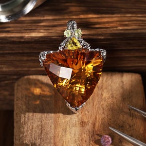 Peut inclure: Une bague avec une grande pierre précieuse triangulaire à facettes de couleur ambre, montée sur une bande en métal argenté texturé. Le sertissage comprend une pierre jaune triangulaire plus petite et deux petites pierres rondes vert pâle.