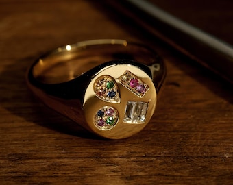 Könnte beinhalten: Ein goldener Siegelring mit einer kreisförmigen Oberfläche. Die Oberfläche zeigt vier Formen, jede mit farbigen Edelsteinen: ein Tropfen, ein Rechteck, ein Kreis und ein Quadrat. Der Ring liegt auf einer Holzoberfläche.
